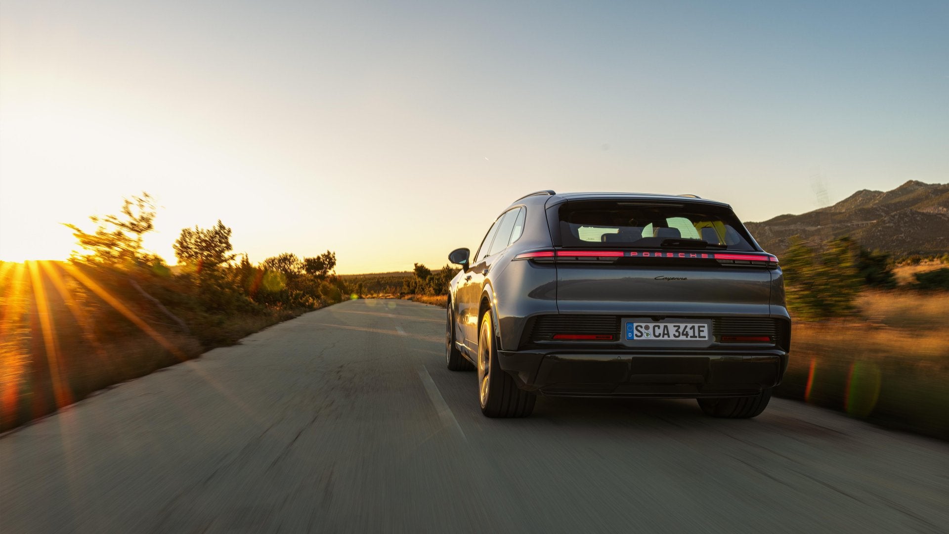 Rear view of the 2026 Porsche Cayenne Turbo Electric driving on a rural road at sunset, showing its full-width LED light bar and sporty stance.